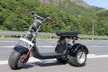 A black three-wheeled electric dirt bike with red accents, parked on an asphalt road with a green forest and rocky mountains in the background.
