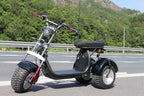 A black three-wheeled electric dirt bike with red accents, parked on an asphalt road with a green forest and rocky mountains in the background.